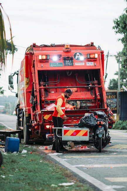 A waste management worker in a high-visibility yellow and red uniform is standing at the rear of a large red refuse collection vehicle, which is actively engaged in rubbish collection on a street. The